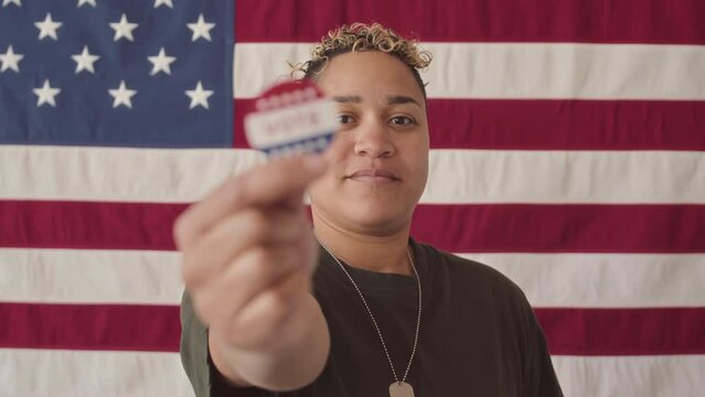 Waist up slowmo portrait of young Biracial military woman standing against US flag at the voting station smiling and showing vote sticker at camera