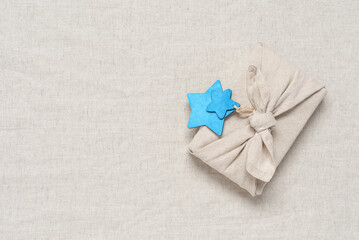 Christmas gift wrapped in fabric with blue wooden stars. Gift in furoshiki style. Beige linen textile background. Zero waste concept. Top view, flat lay. Selective focus.