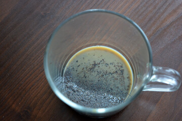 A transparent pleasant glass cup with natural brewed coffee with milk is located on a dark yellow wooden table.