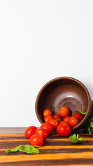 Ripe red cherry tomatoes falling from a clay bowl onto a wooden board with basil leaves on a light white background