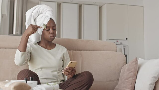 Medium slowmo of young beautiful African American woman with towel on her head doing facial massage with quartz roller while scrolling on smartphone, sitting on sofa in her apartment