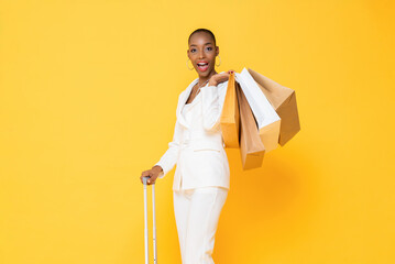 Portrait of excited african american woman with shaved head and mouth open holding shopping bags and suitcase handle against isolated yellow background