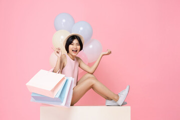 Young smiling Asian woman carrying shopping bags and sitting in pink studio background with balloons