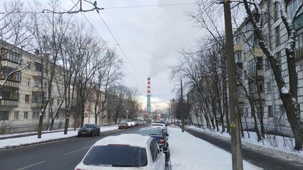 There is smoke coming from the chimney of the power plant. Some cars drive through the city streets, others are parked at the edge of the roadway. After the snowfall, a snow cover formed in the city