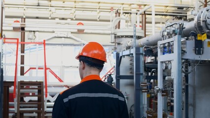 A view from the back of a worker in an orange helmet and overalls walks around his equipment at a chemical plant in the pump shop, checking the condition of the equipment. Factory work.