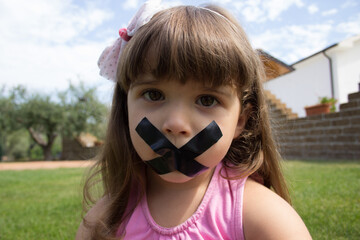 Image of an adorable little girl with a sad expression who has her mouth taped shut.
