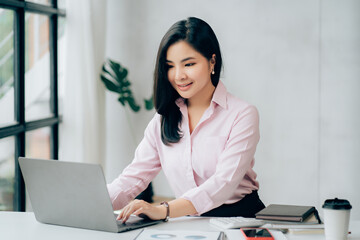 Happy young asian businesswoman sitting on her workplace in the office. Young woman working at laptop in the office