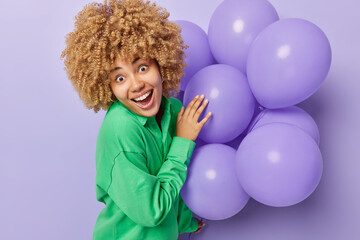 Surprised cheerful European woman with curly blonde hair wears green jumper holds bunch of inflated balloons celebrates university leaving prepares for party isolated over purple background.