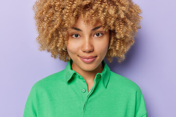 Portrait of good looking European woman with curly bushy hair dressed in green jumper looks directly at camera has calm expression poses against purple background listens information attentively.