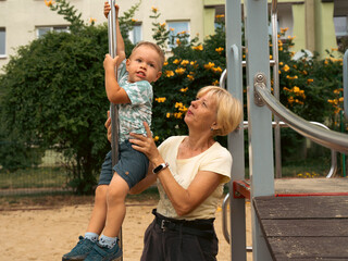 Grandmother supporting and holding her 2 years old grandson at children playground. Grandmother and...