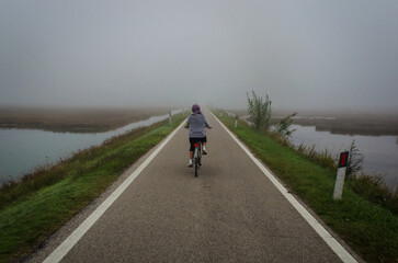Fototapeta premium Una donna va in bicicletta su una strada della laguna nord di Venezia vicino a Lio Piccolo in una giornata di nebbia