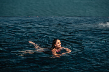 Happy woman in swimsuit swimming in infinity pool against seafront.