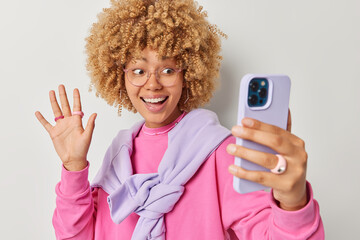 Positive good looking woman with curly blonde hair waves hello at front camera makes video call records blog talks to best friend abroad says hi smiles friendly wears casual outfit poses indoor.