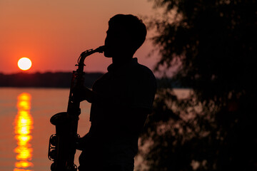 Silhouette of a young guy playing the saxophone in the evening at sunset - obrazy, fototapety, plakaty
