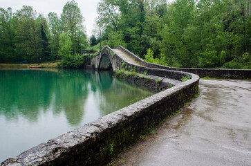 Fototapeta premium Il tipico ponte medievale a schiena di asino detto anche ponte gobbo, si riflette nelle acque di un lago in Garfagnana lungo la Via del Volto Santo, cammino che parte da Pontremoli e arriva a Lucca