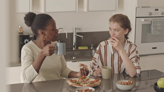 Medium slowmo of delighted young diverse lesbian couple chatting while eating homemade sandwiches at table in modern minimalist style kitchen