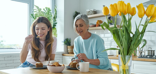 Happy senior mother and her adult daughter enjoying hot drinks and sweet food at the kitchen