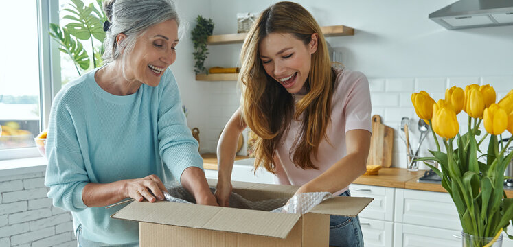 Happy senior woman and her adult daughter unpacking box while standing at the domestic kitchen