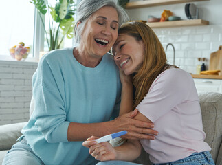 Happy young woman holding pregnancy test and embracing her mother while sitting on the couch together