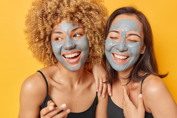 Horizontal shot of happy mixed race two young women smile broadly apply facial mud mask dressed in casual t shirts undergo beauty treatments isolated over yellow background. Skin care procedures