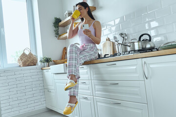 Young woman holding smart phone and drinking coffee while sitting on the kitchen counter