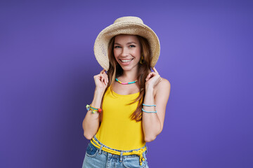 Beautiful young woman adjusting her hat and smiling while standing against purple background