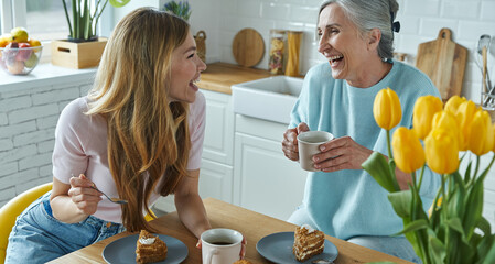 Senior mother and her adult daughter enjoying hot drinks and sweet food at the kitchen