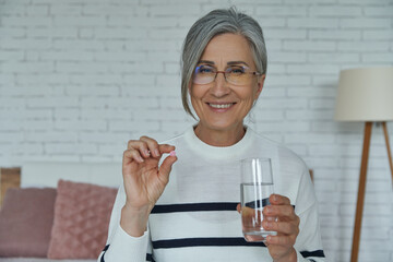 Happy senior woman holding pill and glass with water while sitting on the bed at home