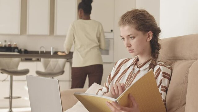 Slowmo of young Caucasian businesswoman sitting on sofa with documents and laptop working from home while young African American housemaid cleaning kitchen in background