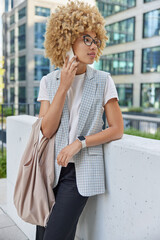 Business and communication concept. Curly haired female managing director speaks on cellphone talks with colleague after work dresed in formal clothes carries bag poses against urban building