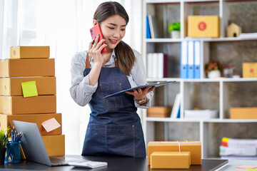 Attractive Asian businesswoman checking items before sending them to customers. Young Asian small business owners use computers. Shipping boxes online market SME e-commerce telemarketing concept