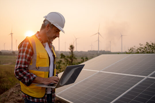 Portrait Of Engineer Wearing Yellow Vest And White Helmet Using A Computer Laptop Checking And Maintenance Of The Solar Panels, Sustainable Energy Concept