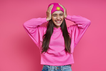 Surprised young woman in hooded shirt holding head in hands against pink background