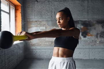Fototapeta na wymiar Concentrated young African woman exercising with kettlebell in gym