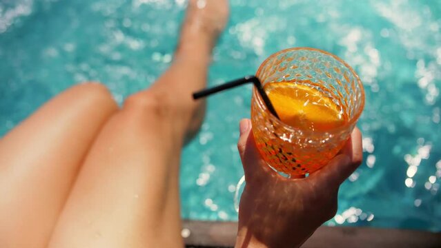 Woman soaking her feet in swimming pool and holding orange cocktail.Young woman drink cocktail near swimming pool.Sexy woman sit near swimming pool relaxing vacation.