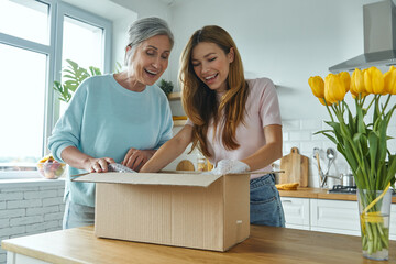 Surprised senior woman and her adult daughter unpacking box while standing at the domestic kitchen