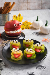 Spooky green kiwi monsters for Halloween. Healthy Fruit Halloween Treats. Halloween party kiwi, strawberry, apple and marshmallow monster on grey stone or concrete table background.  Selective focus.