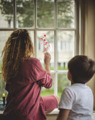 a young woman with curly blond long hair and her son paint pink flowers with paint on the window glass back view
