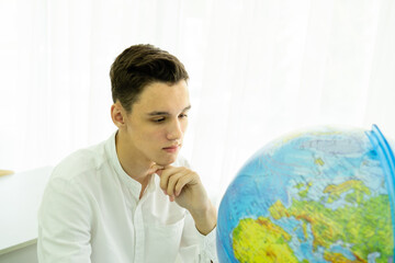 A thoughtful student in a white shirt is sitting at a table in front of a globe. The concept of geography lesson, travel. High quality photo