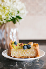 A piece of cheesecake with pistachio and milk cream on the kitchen table near a vase of flowers.