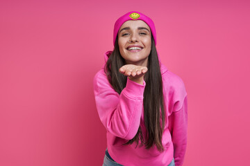 Beautiful young woman in funky hat blowing a kiss against pink background