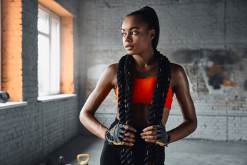 Attractive young African woman carrying battle rope on shoulders while standing in gym