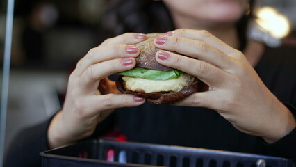 Closeup hand holding hamburger. Young woman eating burger at restaurant. Girl takes a bite of fast food
