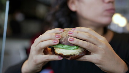 Closeup hand holding hamburger. Young woman eating burger at restaurant. Girl takes a bite of fast food