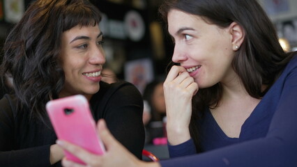 Two young women looking at smartphone device screen smiling. Female friends sharing information watching content online