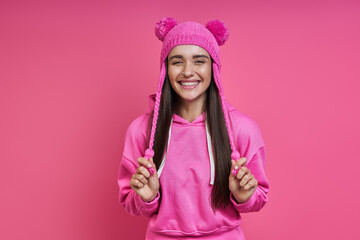 Playful young woman in hooded shirt adjusting her funky hat against pink background