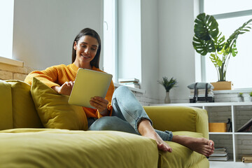 Happy young woman using digital tablet while sitting on the couch at home