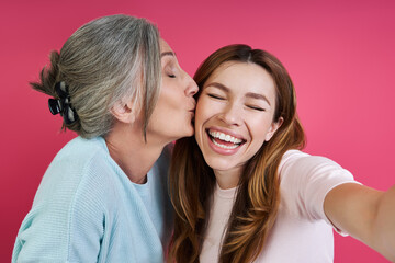 Senior mother kissing her cheerful daughter while making selfie against pink background