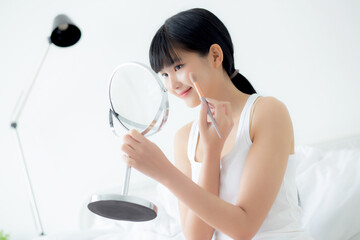 Beautiful young asian woman applying brush on cheek makeup of cosmetic looking mirror on bed, beauty of girl with skin facial and wrinkle in the bedroom, cosmetology for female, skincare concept.