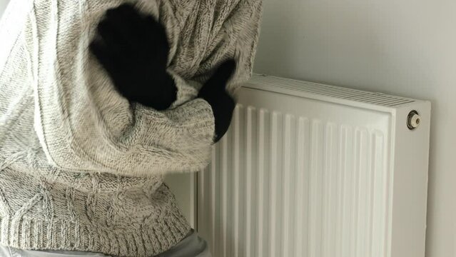 A man in a winter sweater and gloves is shaking from the cold by the radiator, Creative concept, Rising energy prices, Problem with supplying heat to apartments, Inflation attacking household budgets